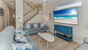 Ein Wohnzimmer mit grauem Sofa, einem weißen Couchtisch, einem blauen Schrank, einem Fernseher, der eine Strandansicht zeigt, und einer Treppe.
