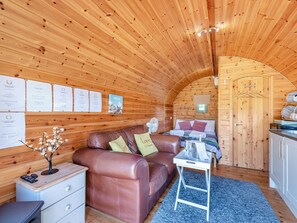 A cozy log cabin interior with a wooden ceiling, a brown leather sofa, a small table with a fan, and a bed with pillows.