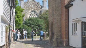 Une rue étroite bordée de bâtiments historiques, dont une église dotée d’une grande rosace et de plusieurs flèches.