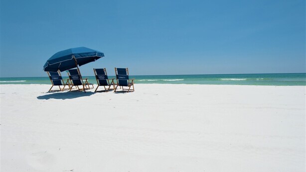 A beach with white sand and a clear blue sky. There are four wooden beach chairs under a blue umbrella.