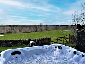 A hot tub with water, surrounded by a stone wall, a wooden gate, and a grassy field with trees and a fence in the background.