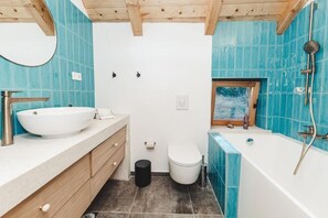 A bathroom with a large white sink, wooden cabinets, a white toilet, a bathtub with a showerhead, and a window with a view of trees. - Villa Terra Meera (Skradin)