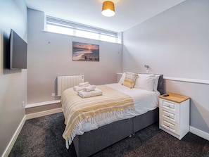 A modern bedroom with a bed, a bedside table, a TV, a radiator, and a window with blinds.