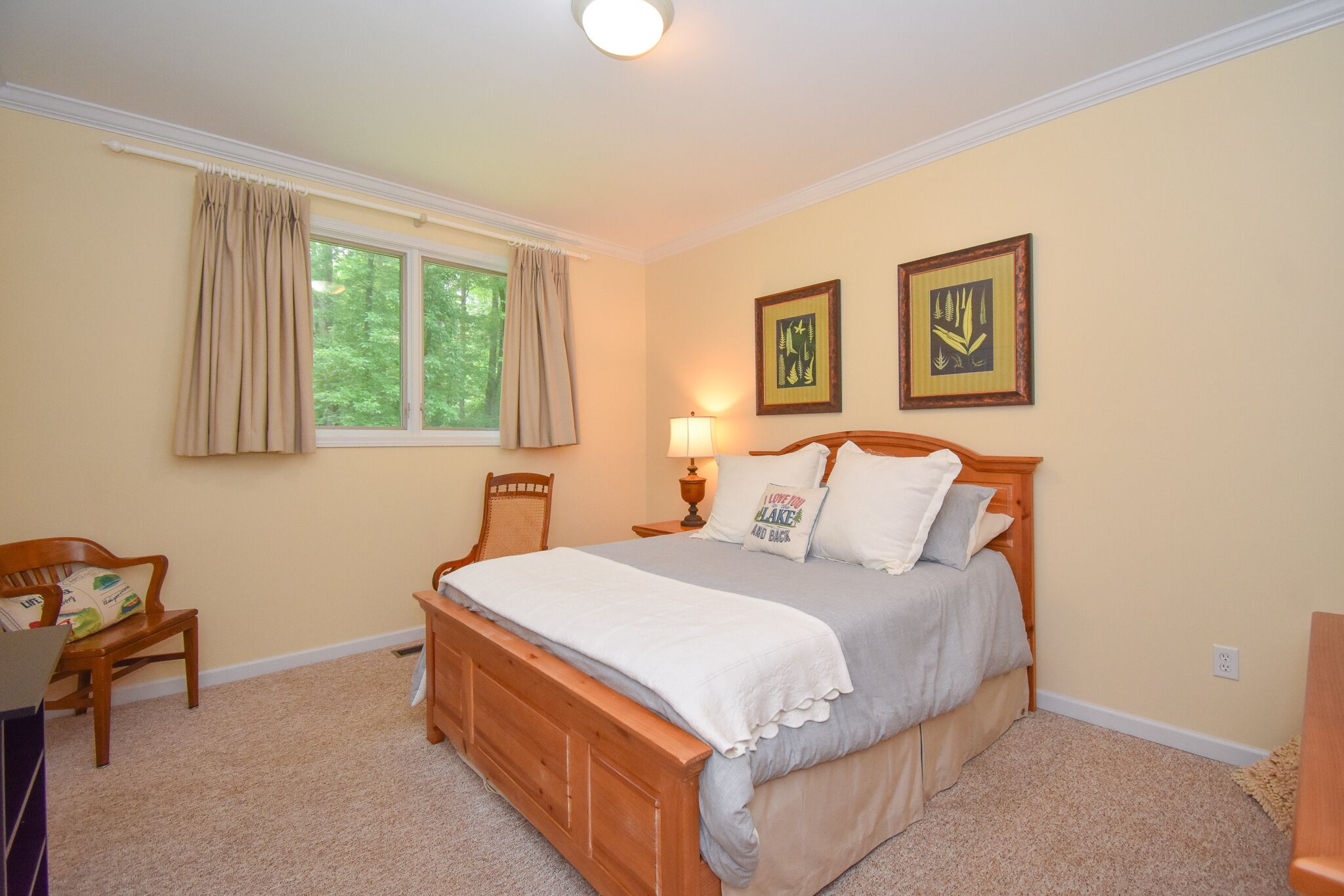 A bedroom with a wooden bed, a nightstand with a lamp, a chair, and two framed pictures on the wall.