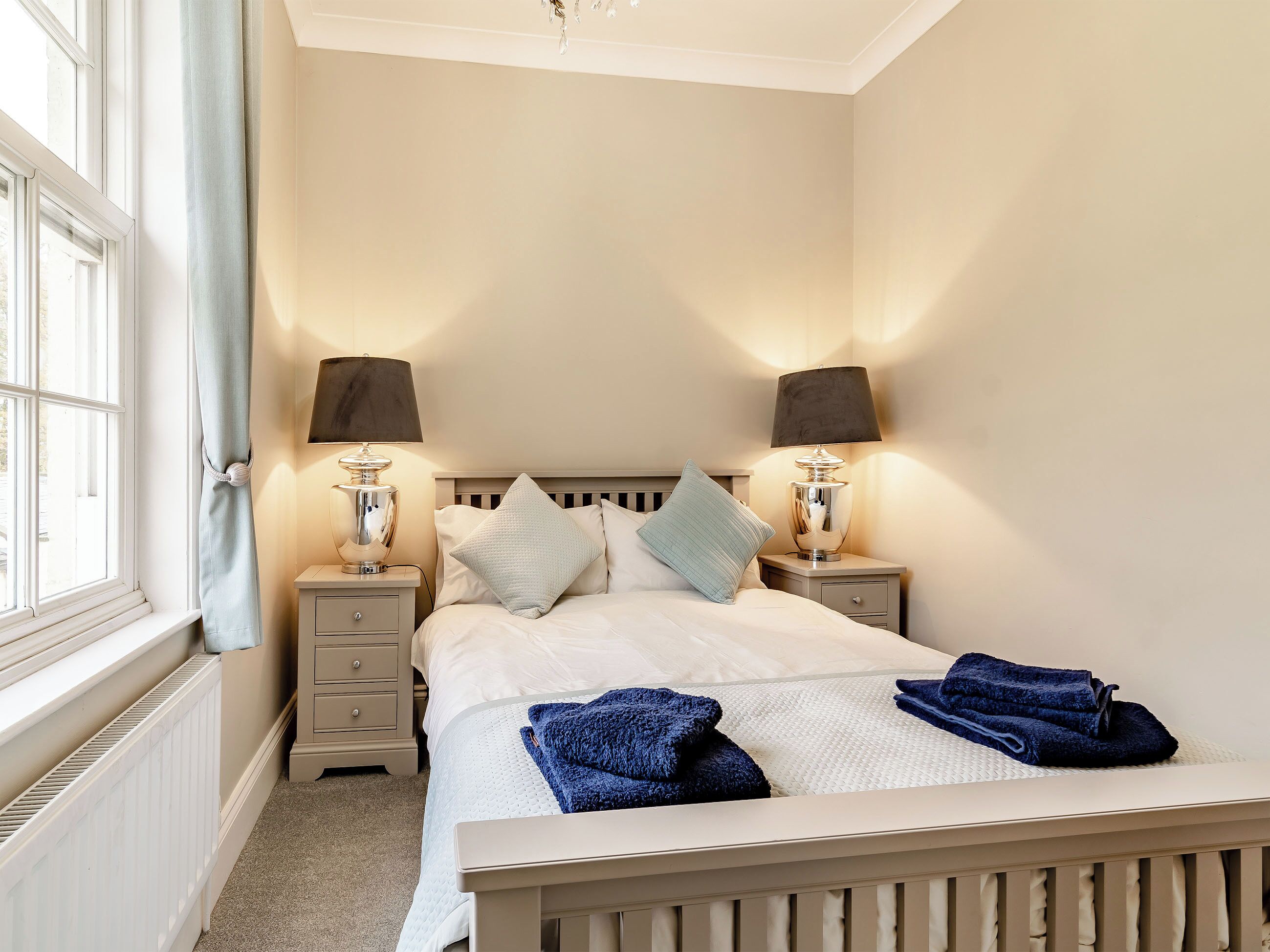Een netjes opgemaakt bed met twee kussens, een nachtkastje met een lamp en een raam met gordijnen.