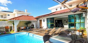 A swimming pool with lounge chairs and a dining area with a glass wall.