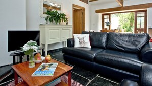 A living room with a black leather sofa, a wooden coffee table with a magazine, a white cabinet with a mirror, and a staircase leading to an upper level. - 2 bedroom accommodation in Chulmleigh (Chulmleigh)