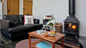 A cozy living room with a black leather sofa, a wooden table with a book and fruit, and a wood-burning stove. - 2 bedroom accommodation in Chulmleigh (Chulmleigh)
