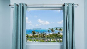 Une fenêtre ornée de rideaux bleu clair, d’où l’on peut admirer une plage bordée de palmiers et l’océan.