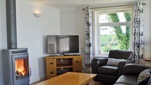 A living room with a wood-burning stove, a television on a wooden stand, a leather sofa, and a large window with curtains.
