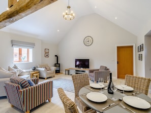 A spacious living area with a high ceiling, a fireplace, a television, and a dining table set for four.
