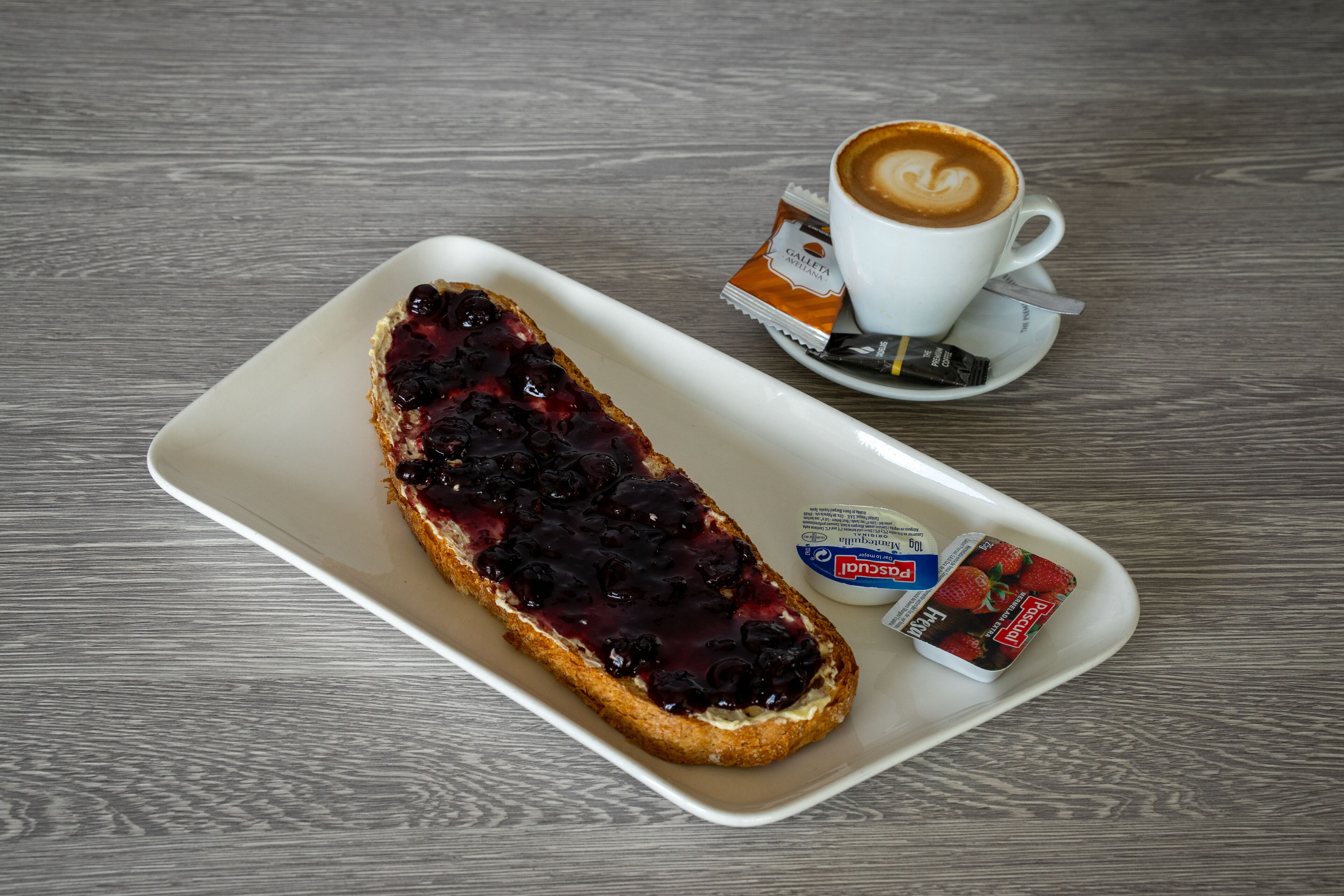 Ein weißer Teller mit Toast, belegt mit Blaubeerkompott, einer Kaffeetasse mit Latte-Art und zwei Päckchen Erdbeermarmelade.