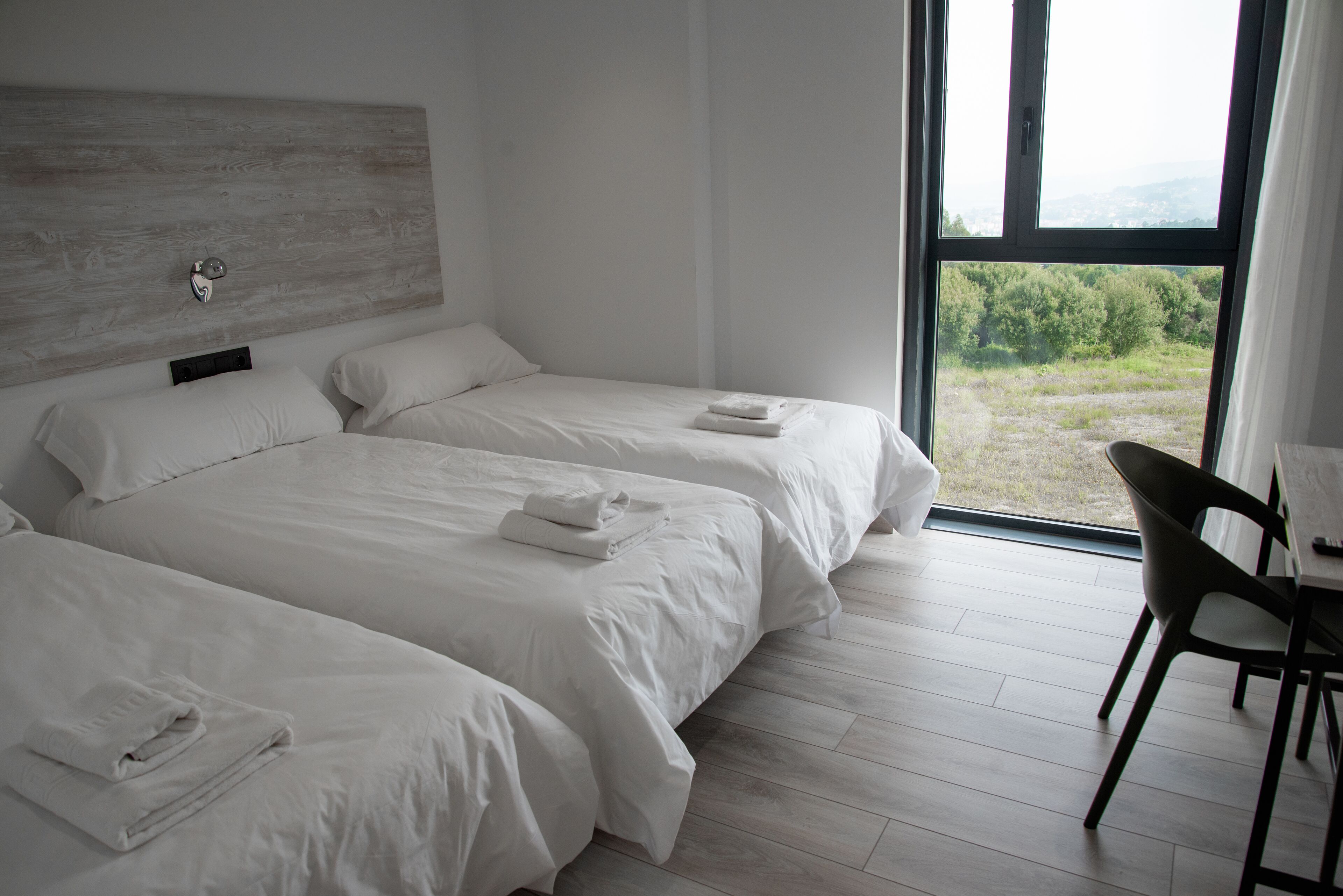 Habitació d'hotel moderna amb dos llits, un panell de capçalera de fusta, una finestra gran amb vista a vegetació i una zona de menjador amb una cadira.