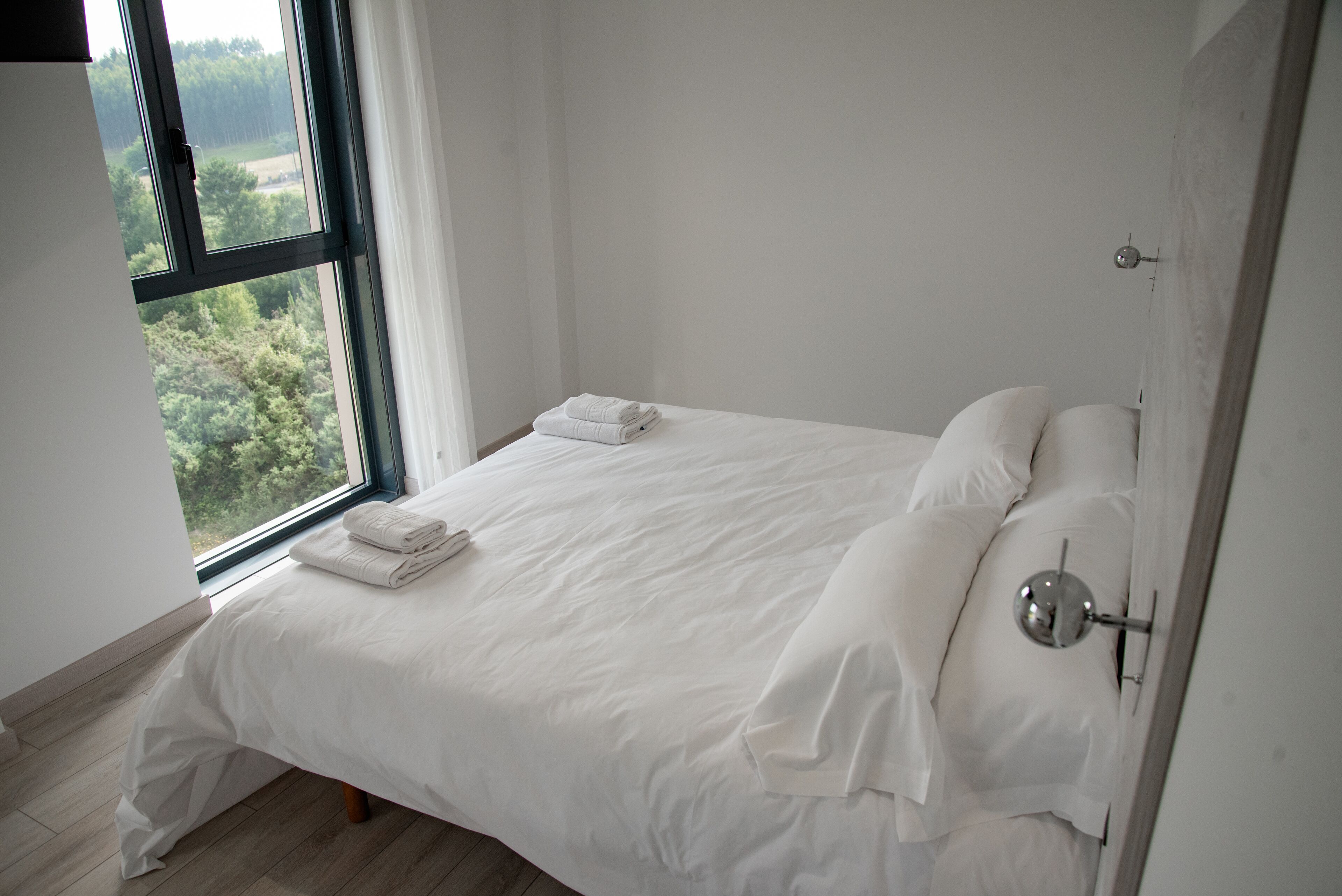 Un dormitorio minimalista con un ventanal grande, una cama bien hecha y una lámpara sencilla de pared.