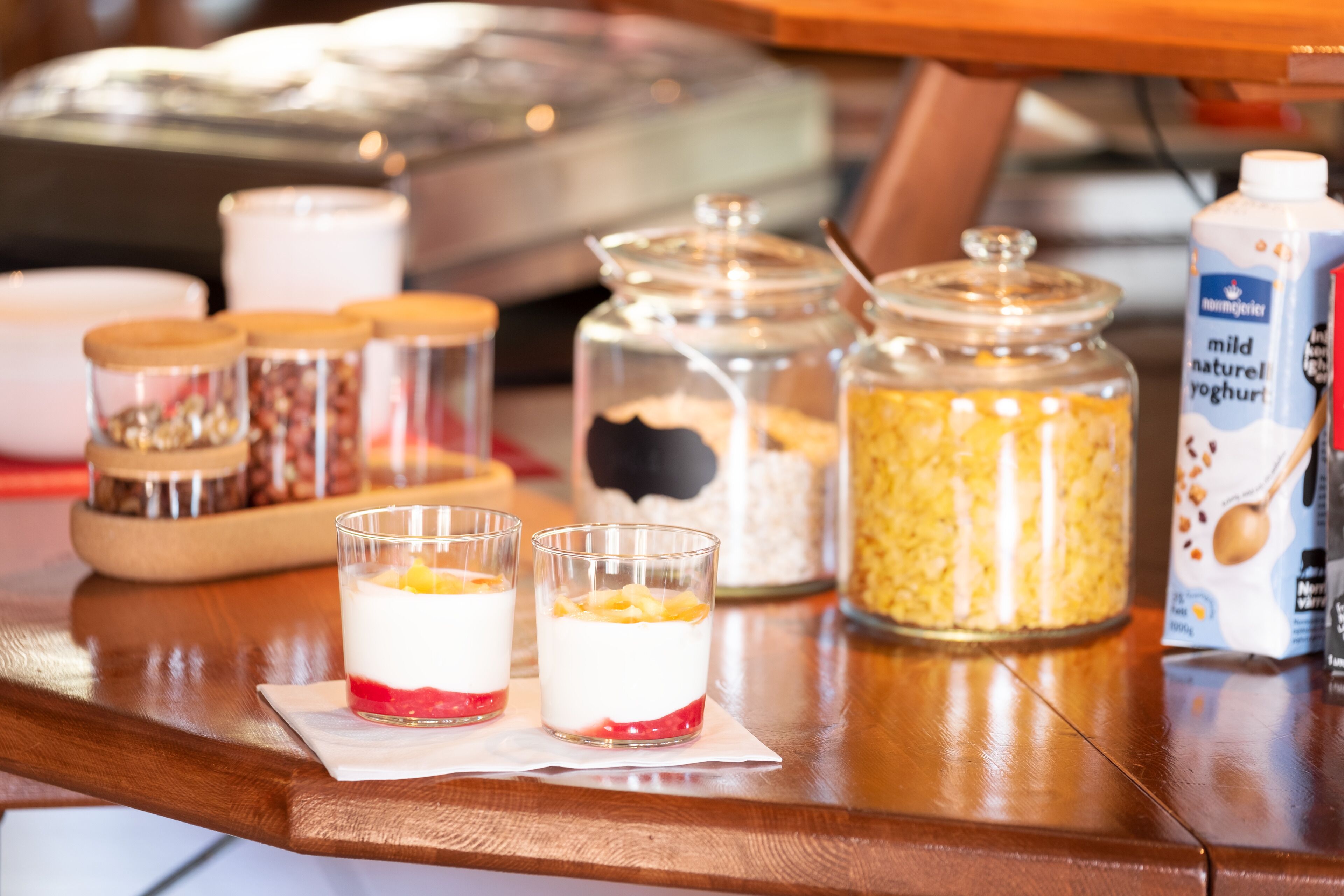 Two glasses of layered dessert on a wooden table with various food containers in the background.