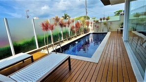 Uma área à beira da piscina com deck de madeira, espreguiçadeiras e vista para um jardim paisagístico com palmeiras.