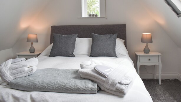 Una cama bien hecha con almohadas, una manta y toallas, dos lámparas de noche, una ventana pequeña y una mesita de noche blanca.