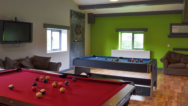 A pool table with balls, a television, and a dartboard in a room with green walls and windows.