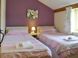 A bedroom with two beds, a nightstand with a lamp, a floral painting on the wall, and a window with curtains.