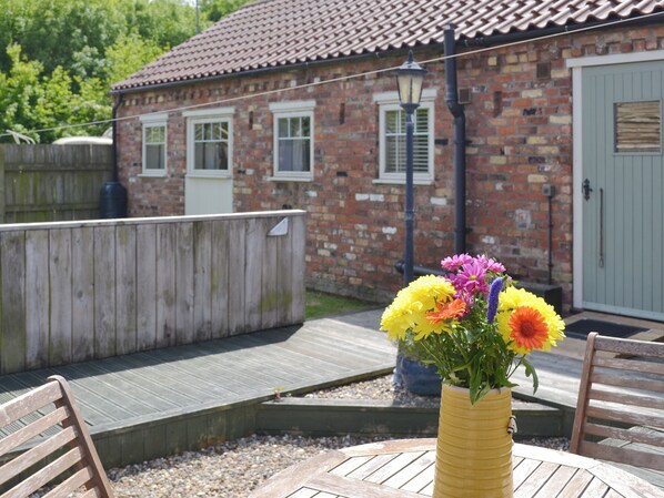 A wooden deck with a bench, a yellow vase with flowers, and a brick building with windows and a door in the background.
