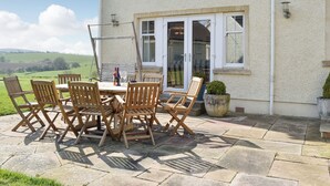 Eine Terrasse im Freien mit Tisch und Stühlen, Blick auf die Landschaft und ein Gebäude mit weißer Fassade und großen Fenstern.