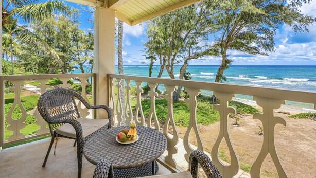 A balcony with wicker furniture, a table with fruit, and a view of the ocean and trees. - View Beachfront, Pool Hot Tub, King Bed (Kapaa)