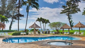 A pool area with lounge chairs and thatched umbrellas, surrounded by palm trees and a view of the ocean. - View Beachfront, Pool Hot Tub, King Bed (Kapaa)