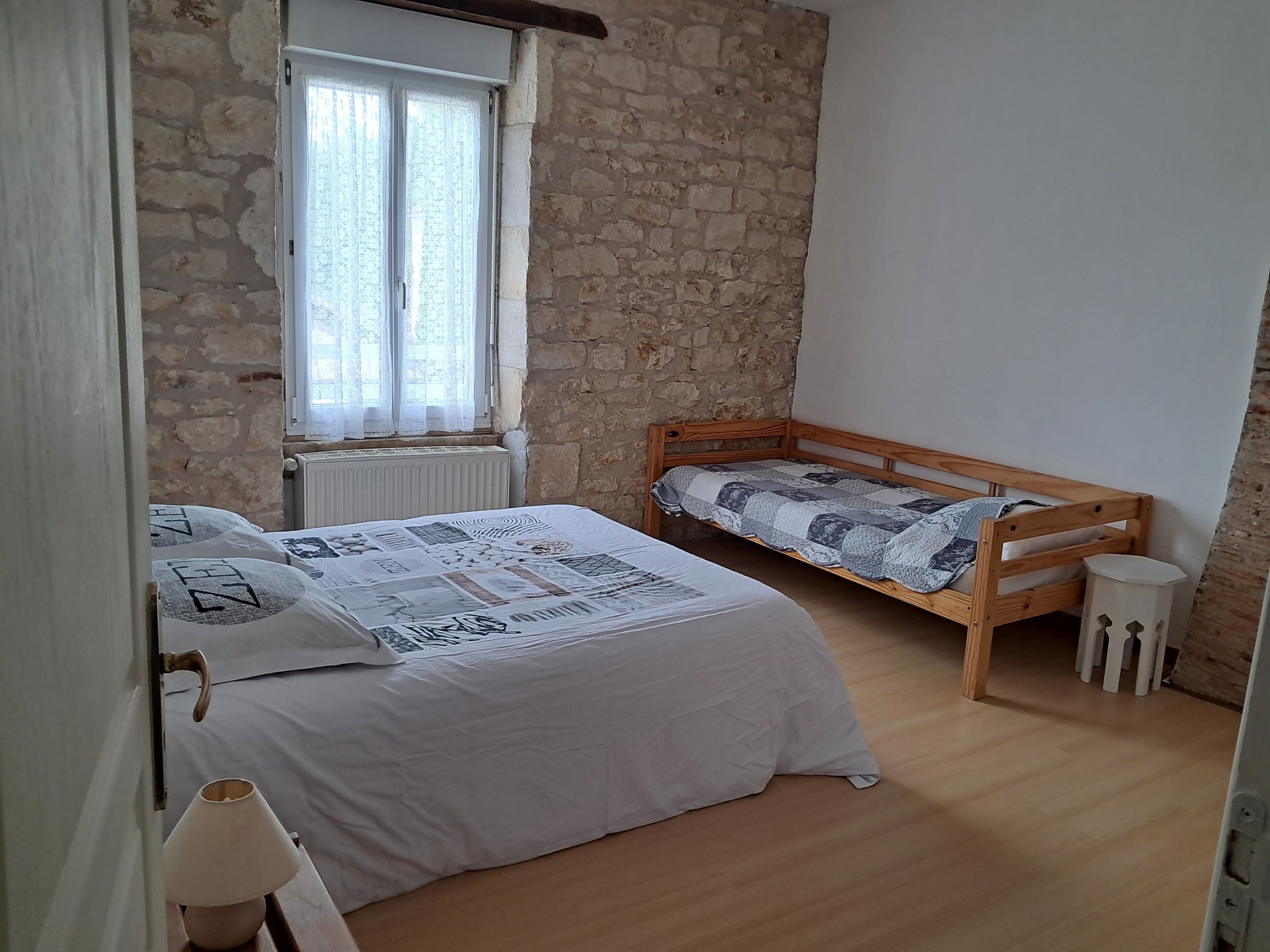 A bedroom with a wooden bed, a smaller bed, a window with curtains, a radiator, and a lamp.
