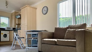 A living room with a beige sofa, a dining table, a washing machine, and a kitchen area with wooden cabinets and a microwave.