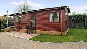 A single-story wooden mobile home with a brick patio, a small garden, and a wooden fence in the background.