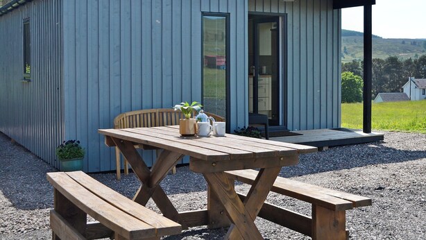 Een laag, eenlaags gebouw met een overdekte veranda, een houten picknicktafel en een bloempot.