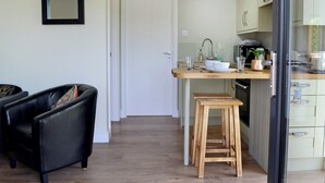 Een compacte keuken met een houten aanrecht, een spoelbak en een klein krukje. Er is een deur die leidt naar een andere kamer en een stoel op de voorgrond.