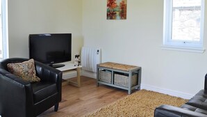 Een woonkamer met een zwarte leren bank, een televisie, een tafeltje met een witte radiator, en een schilderij aan de muur.