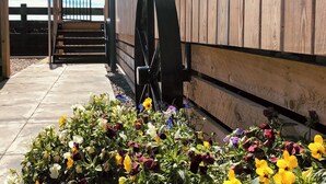 Een houten gebouw met een groot waterrad, een houten hek en een bloembed met diverse bloemen.
