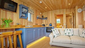 Een gezellig interieur van een chalet met een houten plafond, een keuken met blauwe kastjes, een bank met opvallende kussens en een flatscreen tv aan de muur.
