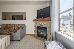 A living room with a stone fireplace, a grey sofa, a wooden coffee table, and a large window with a view of trees and buildings. - Ground Floor Creekside Condo - Pet Friendly, amazing views, near trails (Silver Star Mountain)