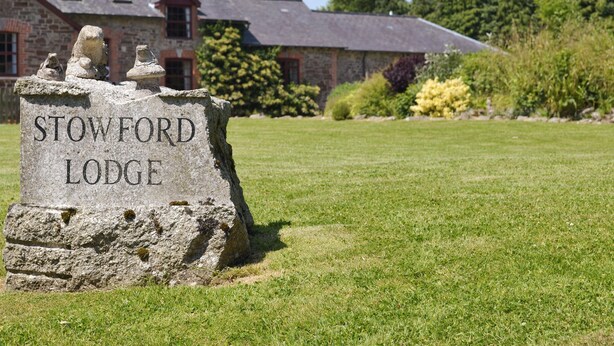 Une plaque en pierre portant l’inscription « Stowford Lodge » est placée devant un bâtiment en pierre doté d’un toit à deux versants et de fenêtres.