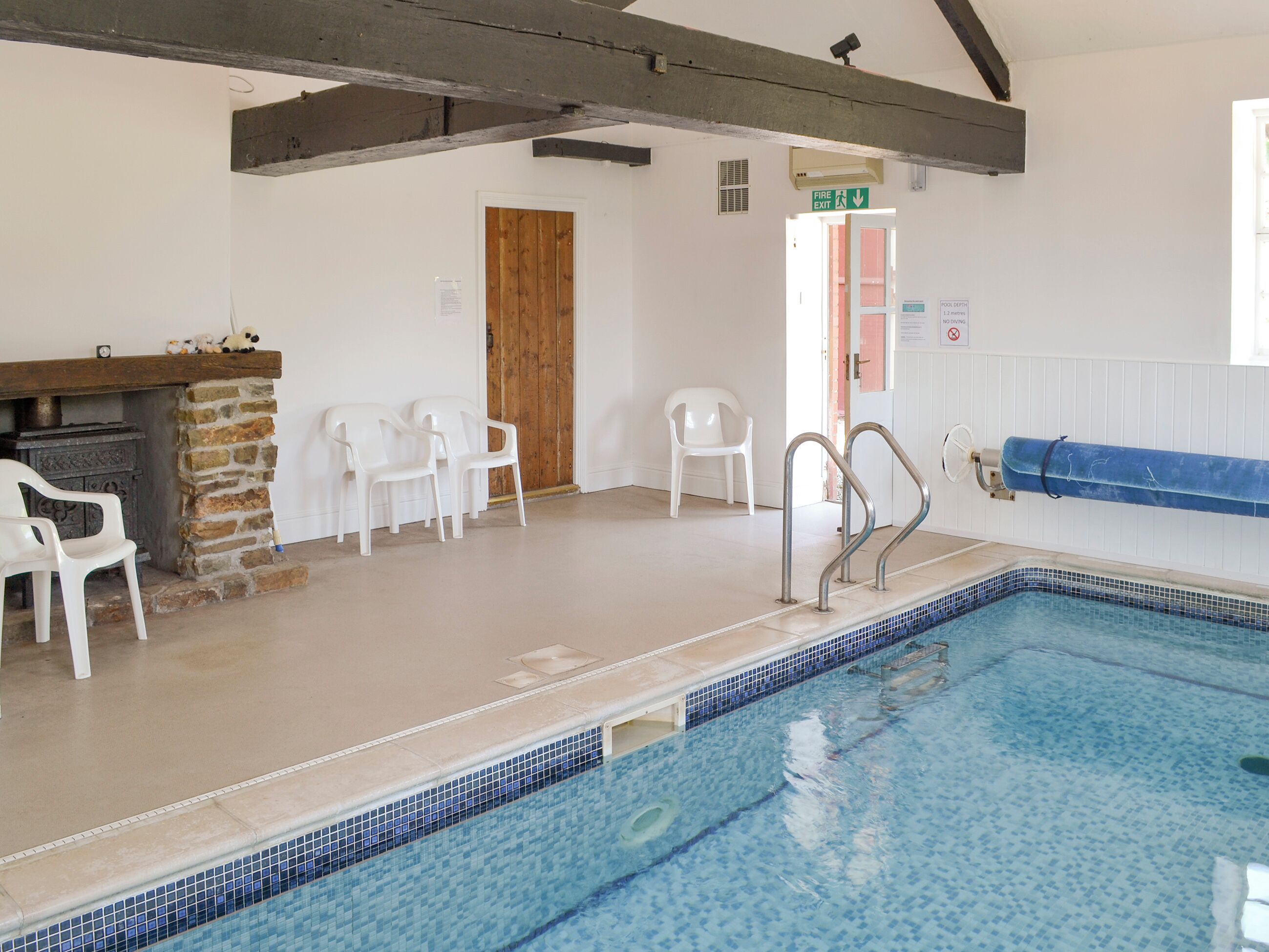 Una piscina con borde de azulejos, chimenea de piedra, sillas de plástico blanco y una puerta de madera.
