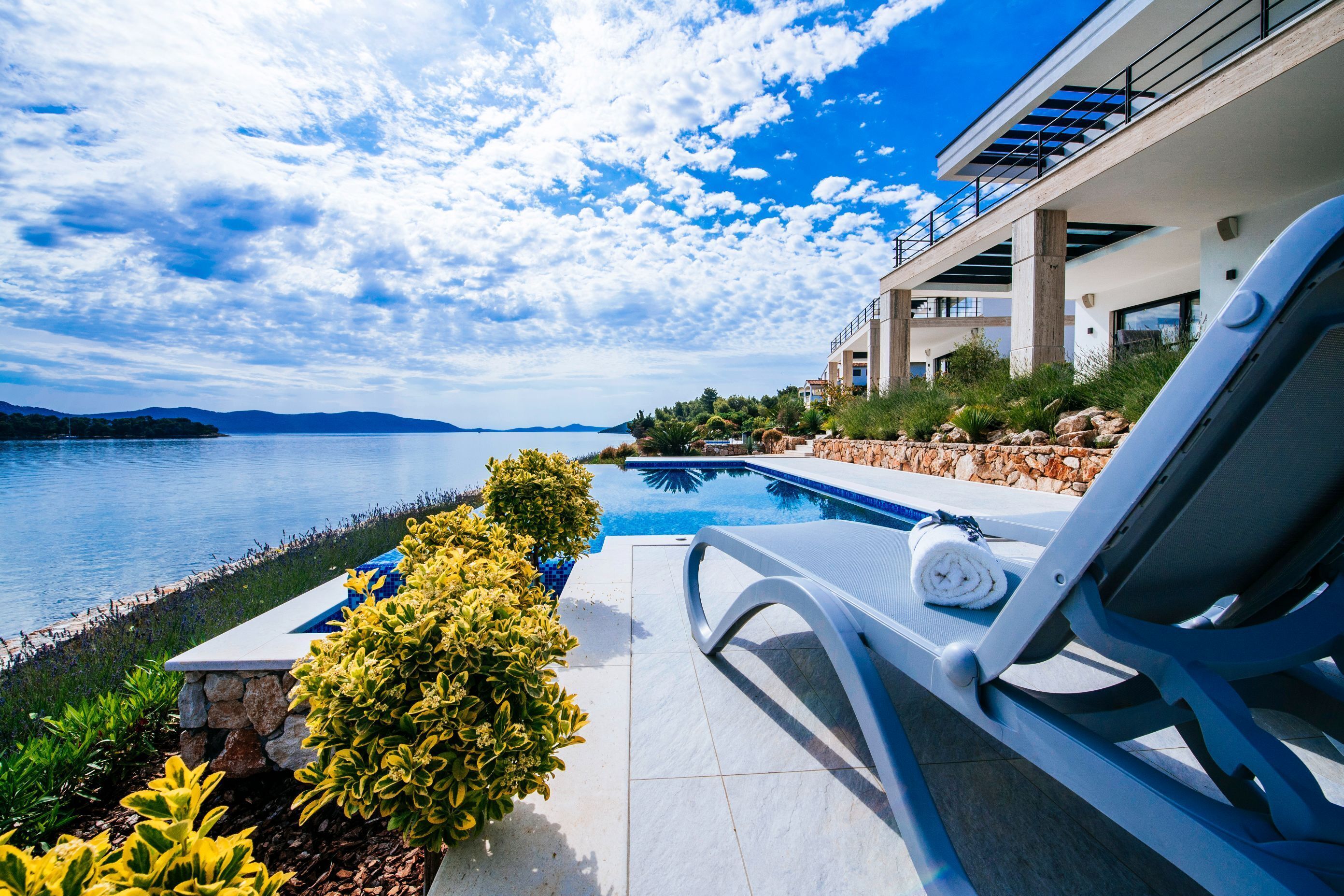 Un área de piscina moderna con vistas al mar y a las montañas, una tumbona y un muro de piedra.