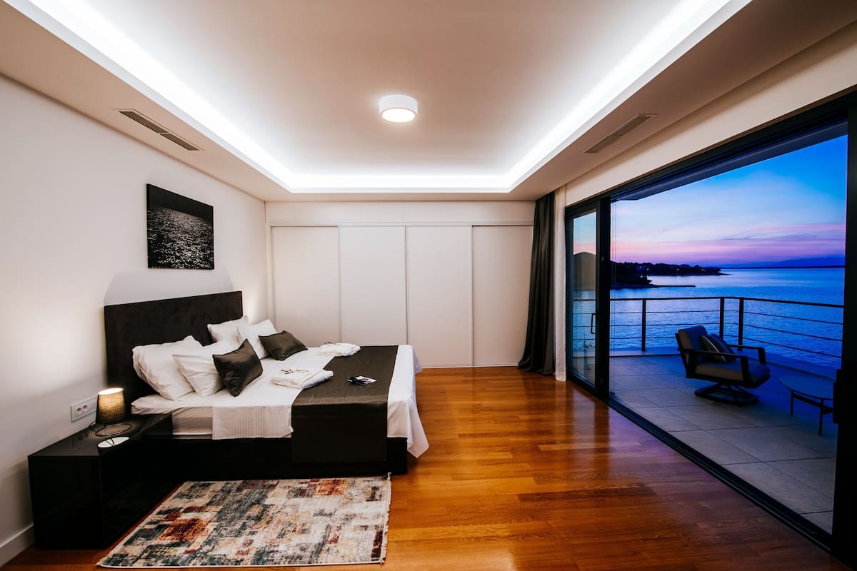 A modern bedroom with a large bed, bedside tables, a rug, and a view of the sea through a glass door.