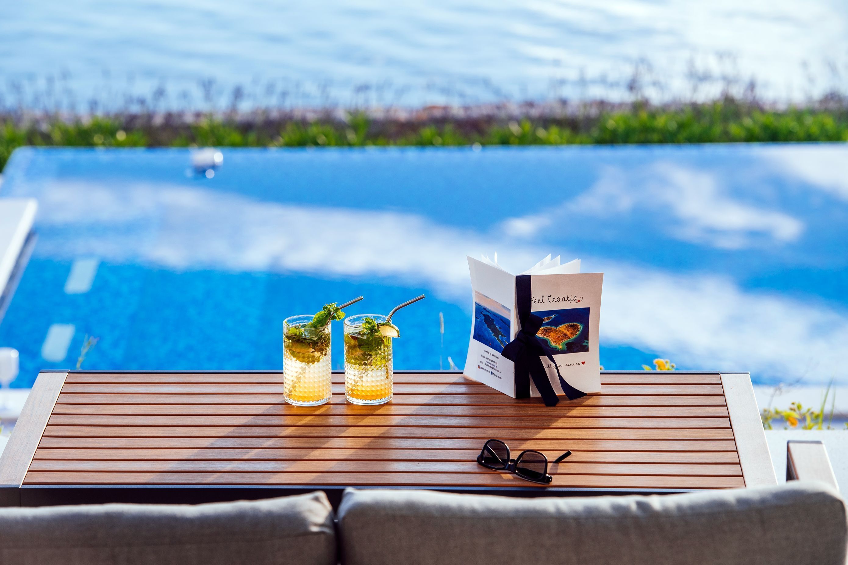 A wooden table with two glasses of a beverage, a brochure, and a pair of sunglasses.