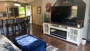 Een moderne woonkamer met een grote flatscreen-tv, een keukenhoek met barstoelen en een knus zithoekje met een blauwe poef.