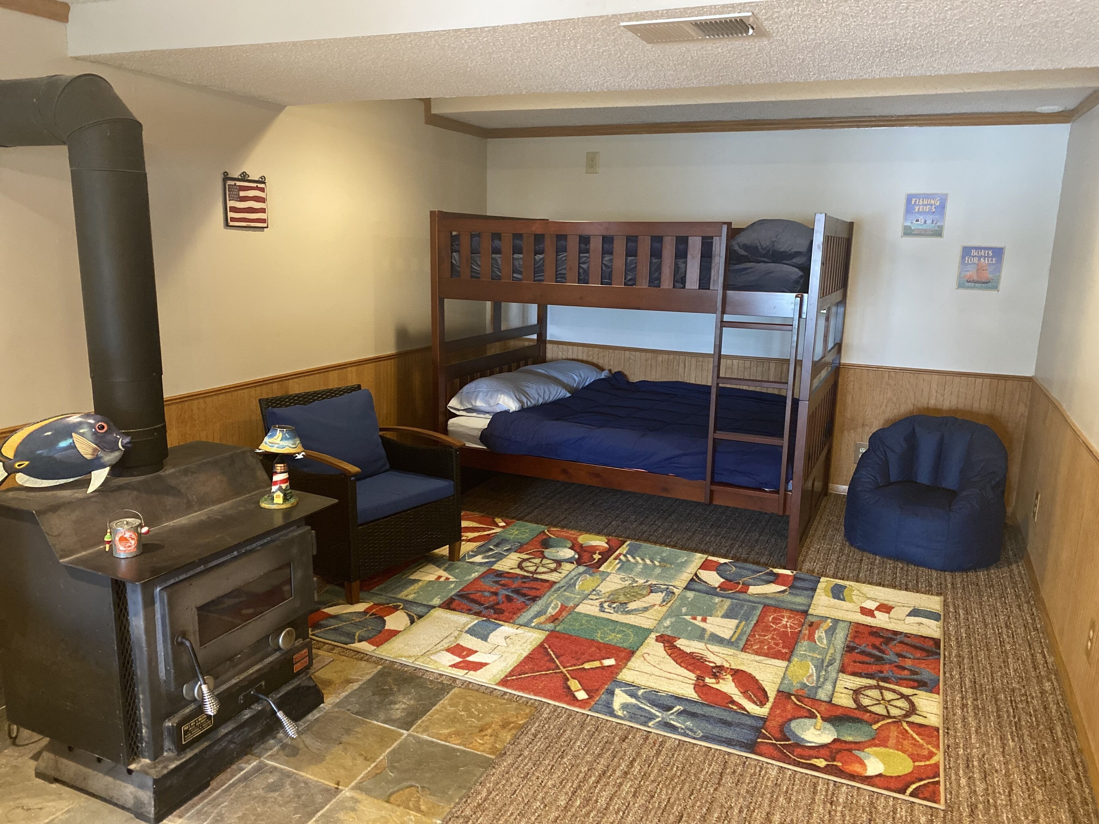 A cozy room with a bunk bed, a small table with a lamp, a chair, a colorful rug, and a wood-burning stove.