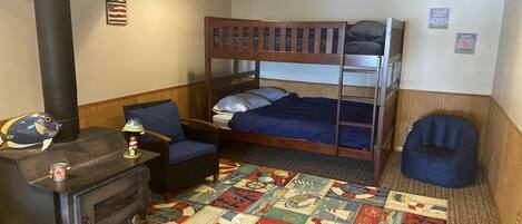 A cozy room with a bunk bed, a small table with a lamp, a chair, a colorful rug, and a wood-burning stove.