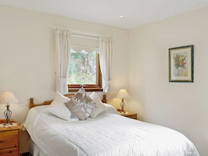A bedroom with a bed, two bedside tables with lamps, a window with curtains, a wall-mounted picture, and a nightstand with a lamp.