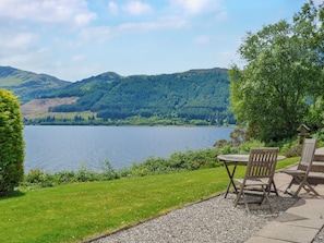 A lakeside view with a table and chairs, surrounded by greenery and hills.