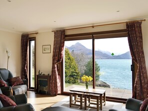 Ein Wohnzimmer mit einer Couch, einem Kamin, einem Esstisch mit Stühlen und Blick auf einen See mit Bergen.