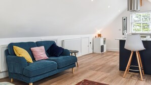 A modern living room with a blue sofa, a small table, and a kitchen area with a window. - 1 bedroom accommodation in Bossiney, near Tintagel (Bossiney, near Tintagel)