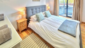 Un dormitorio con una cama grande, dos mesitas de noche, una mesita auxiliar, una ventana con cortinas y un cuadro en la pared.