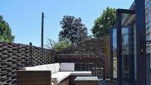 Un sofá de mimbre y un reposapiés sobre una terraza de madera con una puerta de vidrio y un muro tejido.